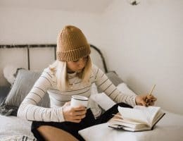 Woman sitting on her bed writing while holding a coffee mug.