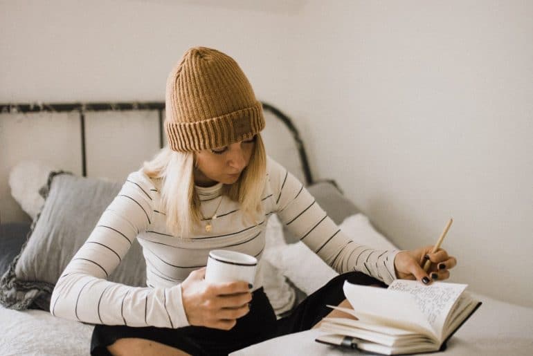 Woman sitting on her bed writing while holding a coffee mug.