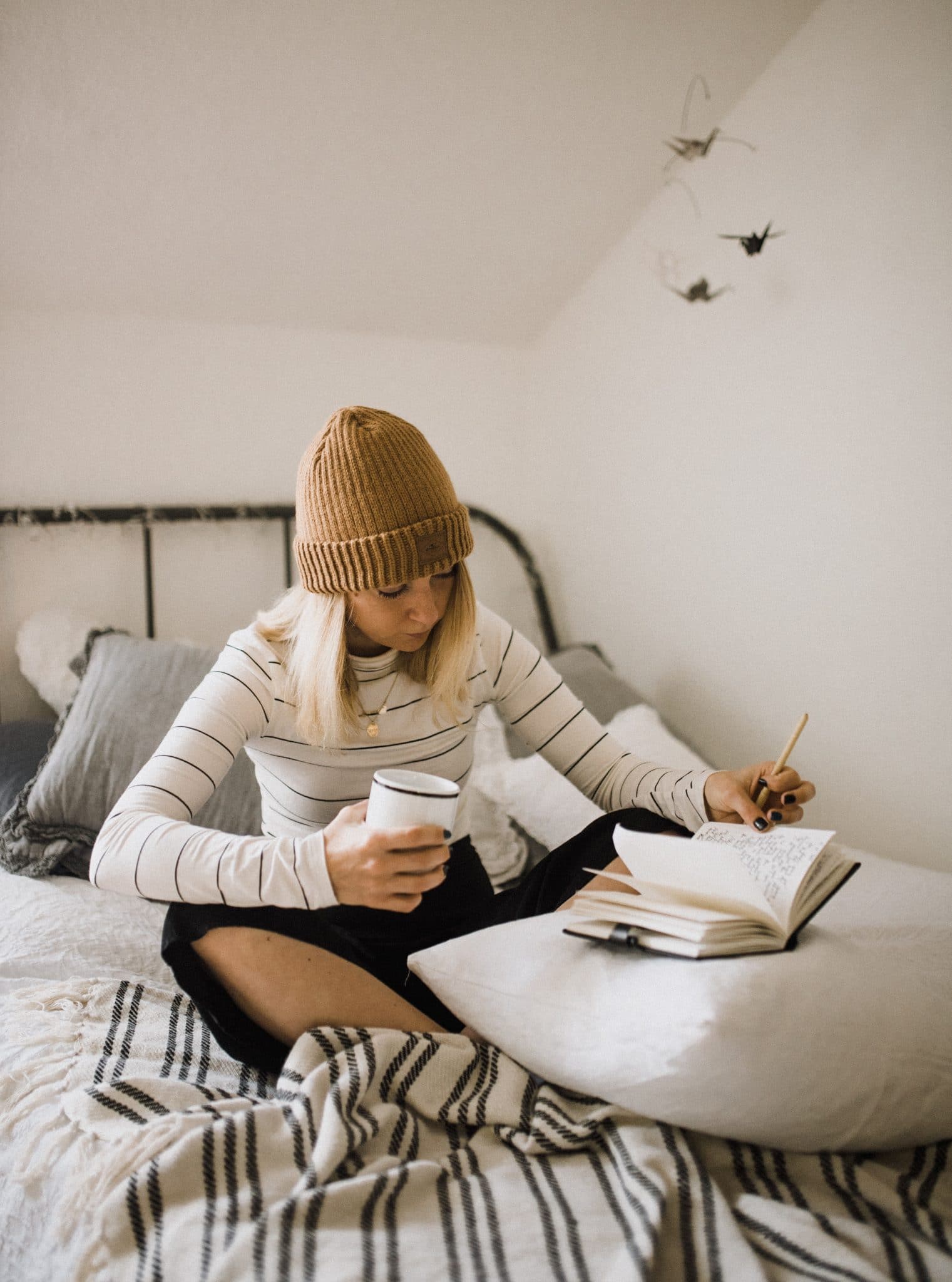 Woman sitting on her bed writing while holding a coffee mug.