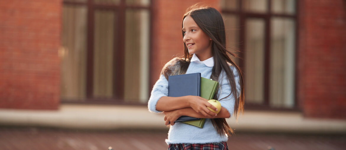 Young school girl in uniform smiling off into the distance with books in hand symbolizing the start of Tachs registration now being open