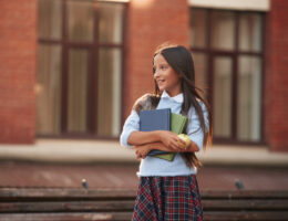 Young school girl in uniform smiling off into the distance with books in hand symbolizing the start of Tachs registration now being open