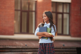 Young school girl in uniform smiling off into the distance with books in hand symbolizing the start of Tachs registration now being open