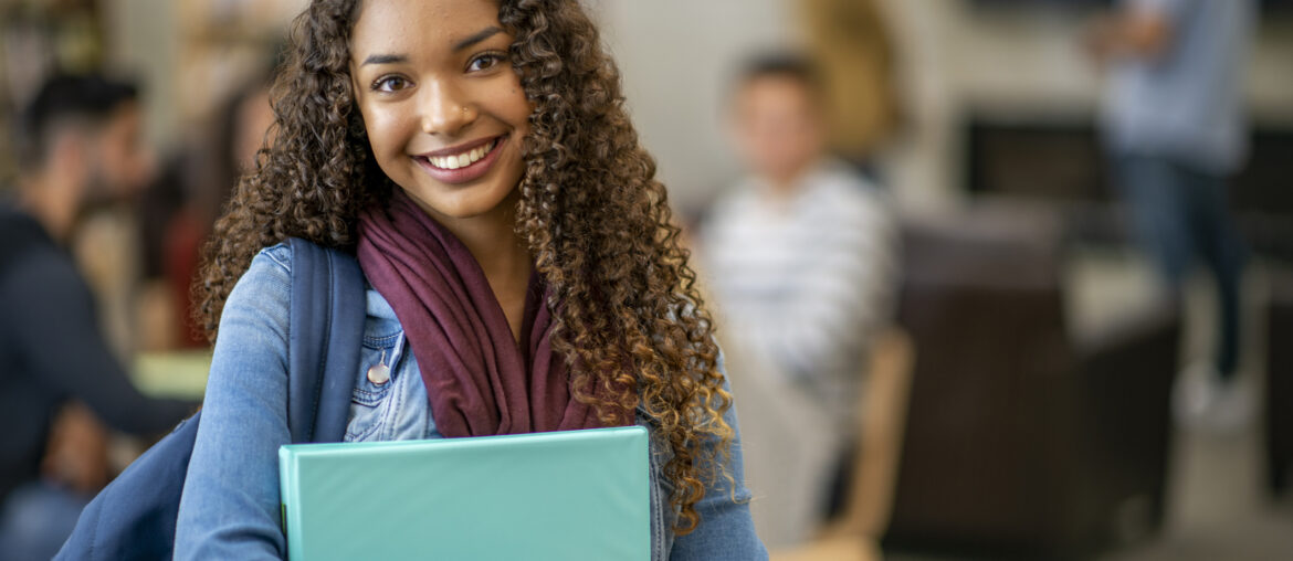 A student smiling at the ACT Test changes while standing in a library with her binders, books, and backpack.