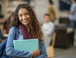 A student smiling at the ACT Test changes while standing in a library with her binders, books, and backpack.