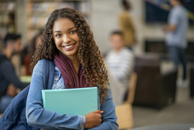 A student smiling at the ACT Test changes while standing in a library with her binders, books, and backpack.