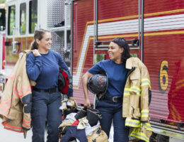 Two female presenting firefighters in front of a fire truck smiling at eachother after passing the Firefighter Exam with Fully Updated prep.