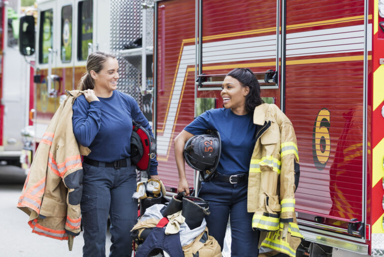 Two female presenting firefighters in front of a fire truck smiling at eachother after passing the Firefighter Exam with Fully Updated prep.