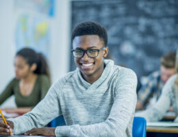 Student with classes in class at a desk preparing to take the ACT, SAT, or CLT for college admissions while smiling.