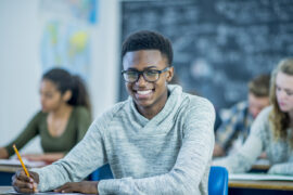 Student with classes in class at a desk preparing to take the ACT, SAT, or CLT for college admissions while smiling.