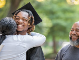 A high school graduate achieving student success by hugging his mom at graduation. His dad is in the background smiling.
