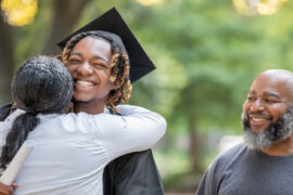 A high school graduate achieving student success by hugging his mom at graduation. His dad is in the background smiling.